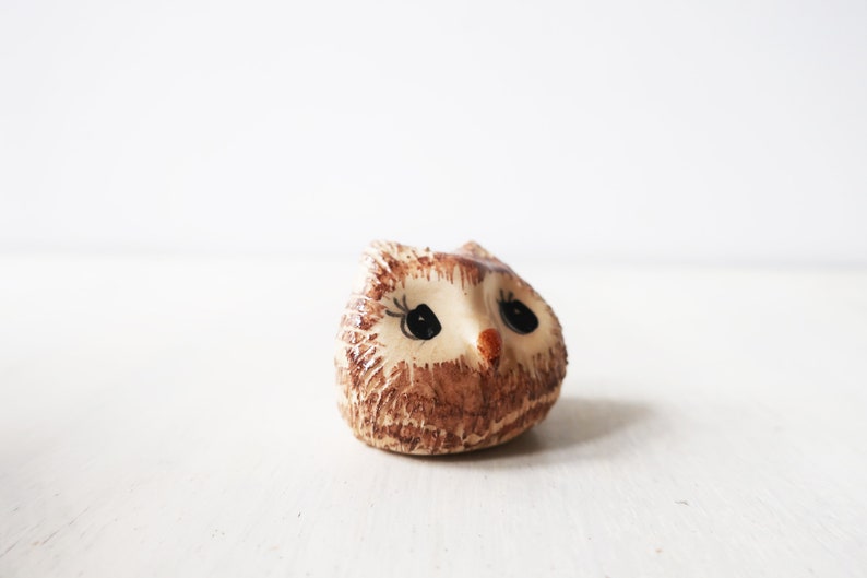 Op de afbeelding: Een kleine keramische uilfiguur met een bruin en beige getextureerd lichaam. De uil heeft grote zwarte ogen, een kleine oranje snavel en delicate wimpers. Decoratief item.