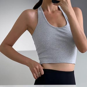 May include: A light gray racerback crop top featuring a scoop neckline and a sleeveless design. The garment is constructed from a soft, heathered fabric and sits at a cropped length, providing a fitted silhouette that ends above the waistline.