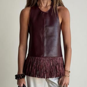May include: A sleeveless burgundy top with a leather-like appearance. The top has a fringe detail at the hem. The model is wearing white trousers and accessories including bracelets and rings.