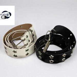 May include: Two rolled belts, one white and one black, each with star-shaped studs. The white belt has a silver buckle and a light brown interior. The black belt also has a silver buckle. Both belts are on a white background.
