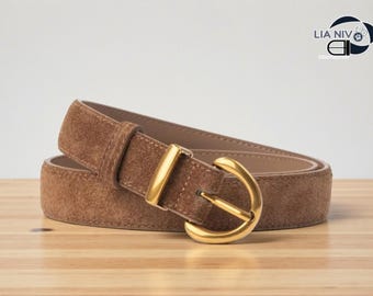 May include: A brown suede belt with a gold-colored buckle. The belt is rolled up on a wooden surface. The buckle is a rounded, D-shaped design. The brand name "LIA NIV" is visible in the top right corner.