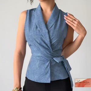 Puede incluir: Un top sin mangas, estilo envolvente, en tejido azul claro. El top presenta un cuello chal y un cierre de lazo en la cintura. El tejido tiene una textura sutil. La parte inferior de la imagen muestra pantalones negros.