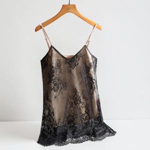 Könnte beinhalten: Ein Camisole mit einer beigen Satin-Basisschicht, die mit komplizierter schwarzer Blumen-Spitze überlagert ist. Das Kleidungsstück hat einen V-Ausschnitt, dünne verstellbare Träger und einen Saum mit Spitzenborte, präsentiert auf einem Holzkleiderbügel.
