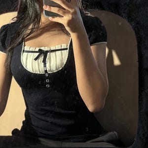 May include: A black short-sleeved top featuring a square neckline with a cream-colored pleated panel. The garment includes a small black ribbon bow at the center of the chest and a vertical row of four clear, circular buttons below the bow.