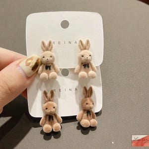 Könnte beinhalten: Vier Plüsch-Hasenohrringe in Braun- und Beigetönen werden auf einer weißen Karte präsentiert. Jeder Hase hat eine kleine schwarze Schleife. Das Wort "REINA" ist auf die Karte gedruckt.