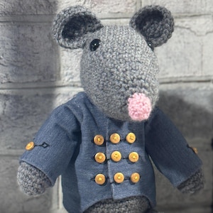 Puede incluir: Un ratón de crochet gris que lleva una chaqueta azul con botones dorados. El ratón tiene la nariz rosa y los ojos negros.