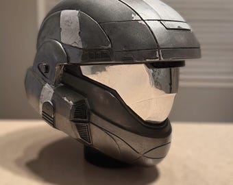 Op de afbeelding: Een metallic grijze helm met zilveren accenten en een reflecterend vizier. De helm heeft een verweerde uitstraling met witte strepen bovenop. Het ontwerp heeft hoekige vormen en ventilatiedetails, wat wijst op een futuristisch of sciencefiction thema.