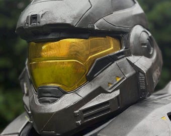 Op de afbeelding: Een metallic grijze integraalhelm met een opvallend goudkleurig vizier. De helm heeft een sculpturaal, hoekig ontwerp met verzonken ventilatieopeningen, zwarte accenten en de letters UNSC in reliëf aan de zijkant, wat een futuristische esthetiek weerspiegelt.