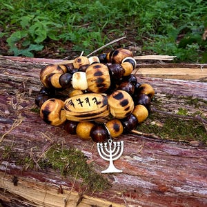 May include: A wooden bead bracelet with a variety of brown and tan beads, including some with dark spots. A silver-colored charm of a menorah hangs from the bracelet. The bracelet rests on a weathered log with green moss.