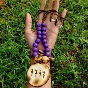 May include: A handcrafted necklace featuring large, round, purple beads strung on a brown cord. The necklace includes a wooden pendant with etched symbols and a wooden bracelet. The background is green grass.