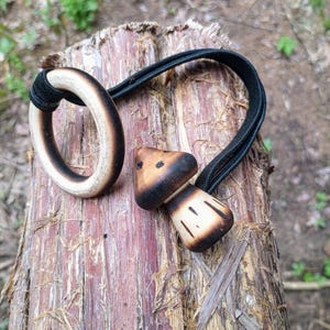 Op de afbeelding: Twee houten haaraccessoires: een ring en een paddenstoelvormige bedel, beide met een verbrand houteffect. De ring is verbonden met een zwarte elastische band, en de paddenstoelbedel is ook bevestigd aan een zwarte elastische band. De accessoires liggen op een boomstam.