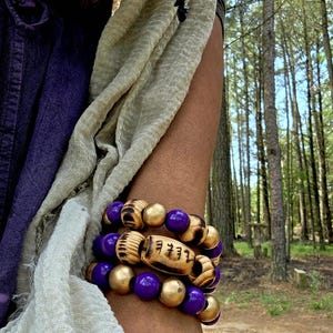 May include: A stack of three bracelets with a forest backdrop. The bracelets feature purple and gold beads, along with wooden beads with etched text. The bracelets are worn on the wrist.
