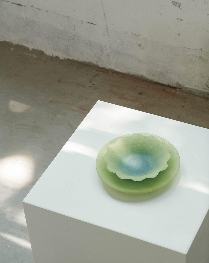 May include: A stack of two glass bowls in shades of green and blue. The top bowl has a scalloped edge and a light blue center. The bowls are sitting on a white surface.
