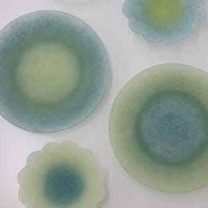 May include: A collection of decorative glass plates in various shapes and sizes. The plates feature a gradient of colors, including shades of blue, green, and yellow. Some plates are round, while others have a scalloped edge. The plates are arranged on a white surface.
