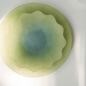 May include: A layered, decorative glass piece with a soft, translucent appearance. The design features a central, scalloped element in shades of green and blue, set within a larger, circular form. The overall effect is organic and serene.
