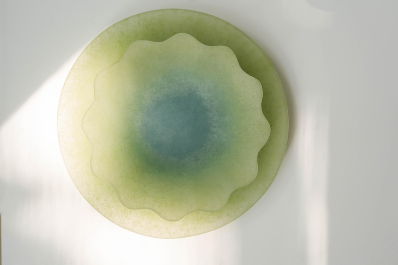 May include: A layered, decorative glass piece with a soft, translucent appearance. The design features a central, scalloped element in shades of green and blue, set within a larger, circular form. The overall effect is organic and serene.