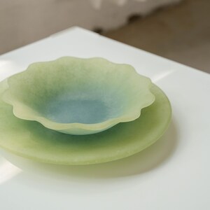 May include: Two-piece decorative glass serving set. The set includes a shallow, scalloped-edge bowl in shades of green and blue, resting on a larger, round plate of a similar color. The pieces are translucent and sit on a white surface.