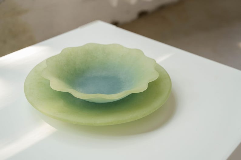 May include: Two-piece decorative glass serving set. The set includes a shallow, scalloped-edge bowl in shades of green and blue, resting on a larger, round plate of a similar color. The pieces are translucent and sit on a white surface.