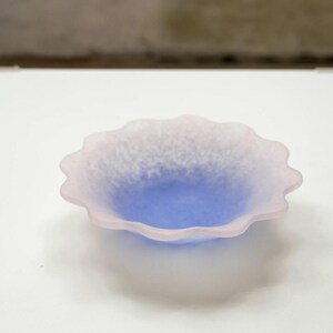May include: A small, decorative bowl with a scalloped edge. The bowl features a gradient of colors, with a light pink rim that fades into a blue center. The bowl is made of a frosted glass material.
