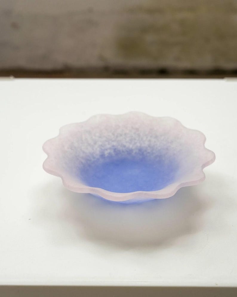 May include: A small, decorative bowl with a scalloped edge. The bowl features a gradient of colors, with a light pink rim that fades into a blue center. The bowl is made of a frosted glass material.
