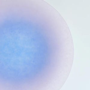 May include: A close-up of a round, light blue and lavender-colored object. The object has a gradient effect, with the blue color concentrated in the center and fading to lavender at the edges. The surface appears slightly textured.