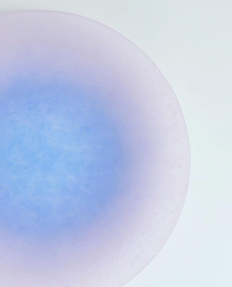 May include: A close-up of a round, light blue and lavender-colored object. The object has a gradient effect, with the blue color concentrated in the center and fading to lavender at the edges. The surface appears slightly textured.