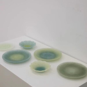May include: A collection of small, decorative glass plates and bowls in shades of green and blue. The plates are round, with a slightly raised edge, and the bowls have a scalloped design. The glass has a translucent quality.