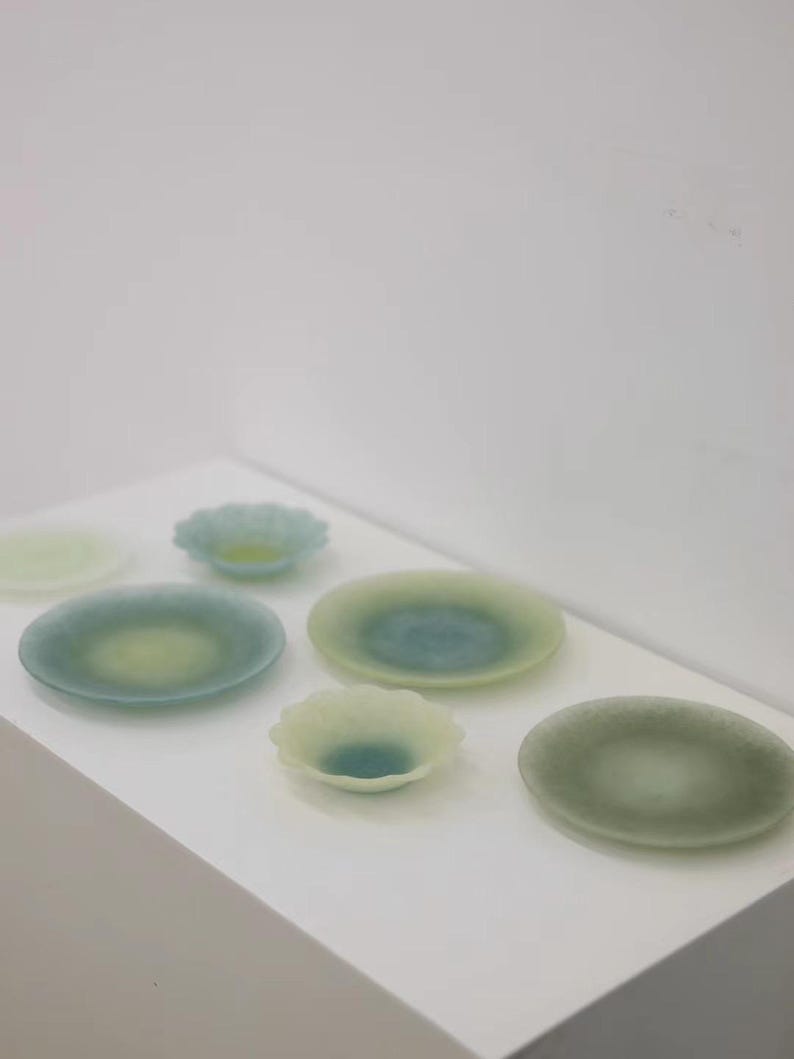 May include: A collection of small, decorative glass plates and bowls in shades of green and blue. The plates are round, with a slightly raised edge, and the bowls have a scalloped design. The glass has a translucent quality.