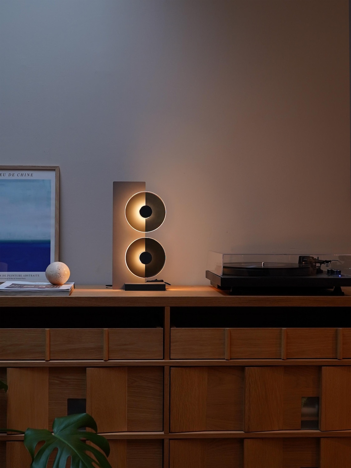 May include: A modern, minimalist lamp with two circular light fixtures. The lamp is on a wooden shelf with a record player and a framed poster. The lamp is made of a light brown material and has a black base. The light fixtures are made of a dark gray material and have a white inner circle. The light fixtures are illuminated with a warm, white light. The record player is black and has a clear lid. The framed poster is white and has a blue and white image of a beach scene. The poster has the text 'DEKE DE CHINE' in black letters at the top.