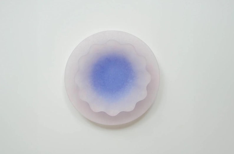 May include: A round, layered object with a soft, blurred appearance. The outer layer is a pale pink, while the center features a vibrant blue hue. The edges of the inner layer have a scalloped design. The object is set against a white background.