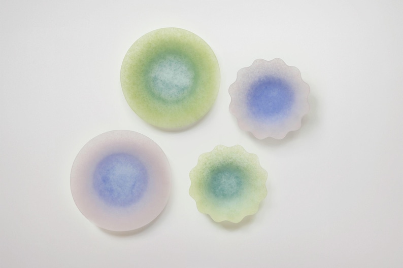 May include: Four decorative glass pieces in shades of green, blue, and purple. Two are round, and two have scalloped edges. The colors blend from the edges to the center, creating a soft, ethereal effect. The pieces are arranged on a white surface.