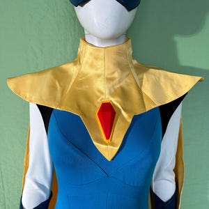 May include: A superhero costume on a mannequin. The costume features a blue bodysuit, a gold collar, and a red gem. The sleeves are white with navy blue accents. A navy blue mask completes the look.