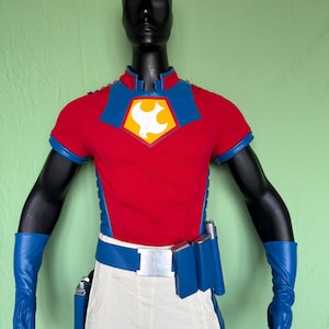 Puede incluir: Un disfraz de superhéroe rojo y azul exhibido en un maniquí negro. El disfraz incluye una camiseta roja de manga corta con cuello y detalles azules, pantalones blancos, un cinturón azul y guantes azules. Un emblema de paloma blanca está en el pecho.