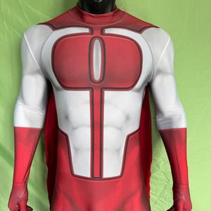 Puede incluir: Un disfraz de superhéroe rojo y blanco en un maniquí. El disfraz presenta un torso rojo con un emblema blanco, brazos blancos y guantes rojos. Una capa roja es visible en la parte posterior. El maniquí tiene una cabeza negra.