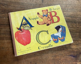 May include: A vintage children's book with a yellow cover featuring the letters A, B, and C. The letter A is illustrated with a red apple, the letter B is illustrated with a teddy bear, and the letter C is illustrated with a baby carriage. The book's title is "A-apple, B-bear, C-cradle".