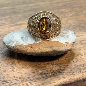 May include: A gold-toned class ring featuring an oval-cut amber-colored stone at the center. The bezel is engraved with the text COLLEGE OF NOTRE DAME and the year 1989. The sides of the ring display intricate embossed metal detailing.