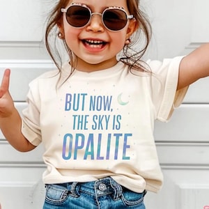 May include: A smiling child wearing sunglasses and a cream-colored t-shirt with the text "BUT NOW, THE SKY IS OPALITE" in blue and purple. The child is wearing blue jeans and making a peace sign with their fingers.