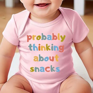 May include: A light pink baby onesie with the text "probably thinking about snacks" in colorful letters. The onesie is short-sleeved and has a simple, classic design, perfect for a baby.