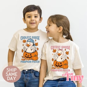 May include: Two children wearing cream-colored t-shirts with goose-themed graphics. One shirt says "Coolest Goose at the Pumpkin Patch" with a goose in a pumpkin. The other says "Cutest Goose at the Pumpkin Patch" with a goose in a pumpkin.