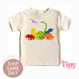 May include: Cream-colored t-shirt featuring a colorful dinosaur graphic. The design includes a red Triceratops, yellow Tyrannosaurus Rex, green Brachiosaurus, blue Stegosaurus, and orange Stegosaurus. A purple Pterodactyl is also included. The shirt has the text "Tiny Trends Tee".