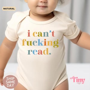 May include: A cream-coloured baby onesie with the text "i can't fucking read" in colourful letters. The onesie has short sleeves and a round neckline. The word "NATURAL" is printed in a small box in the upper left corner.