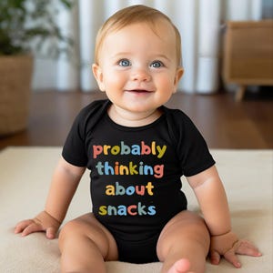 May include: A smiling baby wearing a black onesie with the text "probably thinking about snacks" in colorful letters. The baby has blue eyes and fair skin, sitting on a light-colored surface.