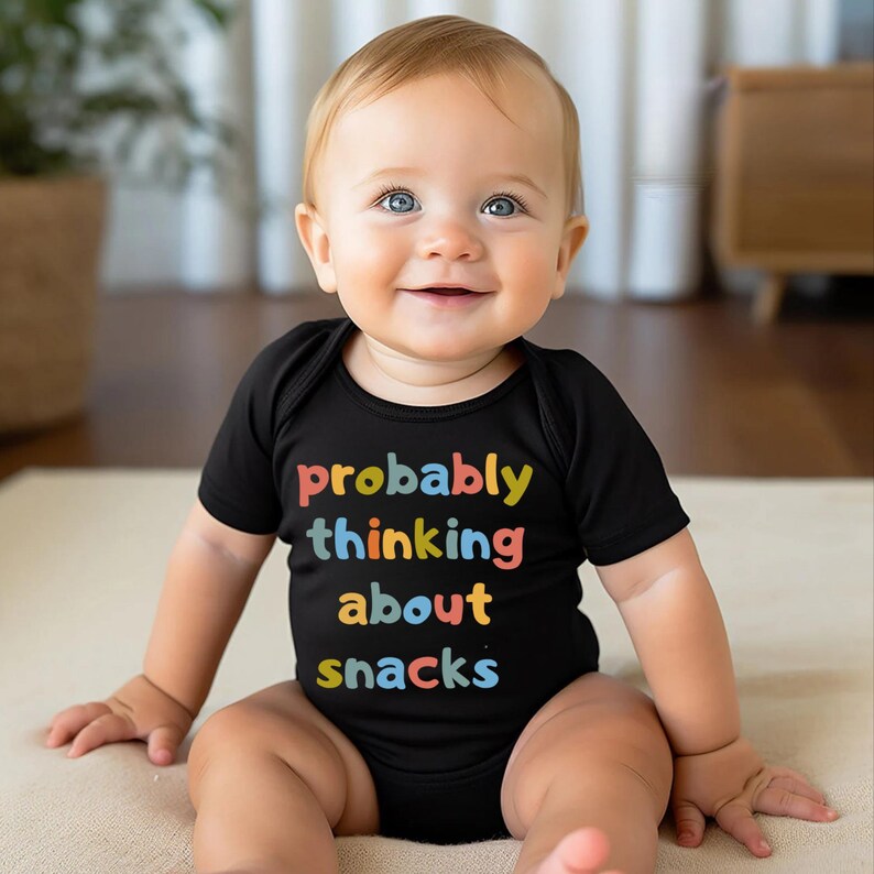 May include: A smiling baby wearing a black onesie with the text "probably thinking about snacks" in colorful letters. The baby has blue eyes and fair skin, sitting on a light-colored surface.