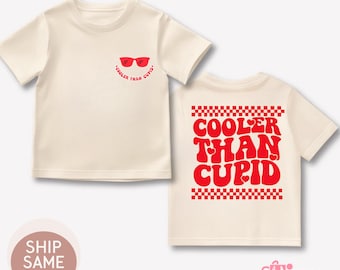 May include: Cream-colored t-shirt with the text "Cooler Than Cupid" in red. The front of the shirt features red sunglasses and the same phrase. The back of the shirt has the phrase in a larger font with a checkered pattern.