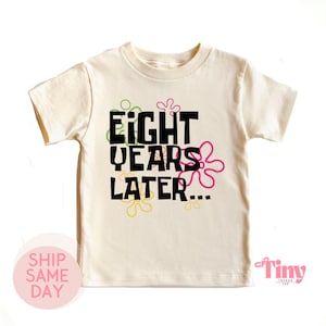 May include: A cream colored t-shirt with a black graphic that says "Eight Years Later..." surrounded by colorful flowers.