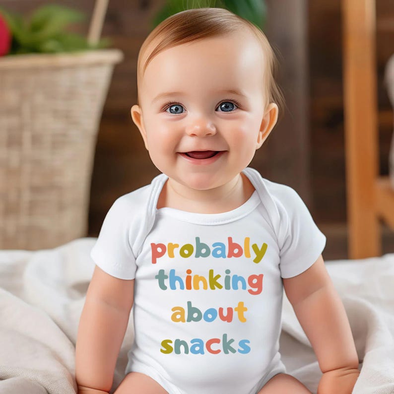 May include: A smiling baby wearing a white onesie with the text "probably thinking about snacks" in colorful letters. The baby has blue eyes and light brown hair.