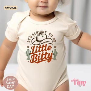 Peut inclure: Un body de bébé crème avec le texte "It's Alright to be Little Bitty" en marron et blanc. Le motif comprend un chapeau de cowboy et des cactus. Le body est à manches courtes.
