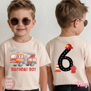 May include: A light beige t-shirt featuring a red fire truck graphic with the text BIRTHDAY BOY on the front. The back displays a large black number 6 shaped like a road, topped with a red firefighter helmet, next to a traffic cone and hydrant.