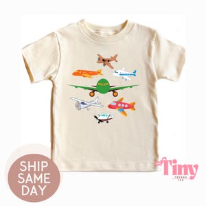 May include: Cream-colored t-shirt featuring a colorful airplane graphic. The design includes various cartoon airplanes in orange, green, red, white, and brown. The shirt has short sleeves and a crew neck.