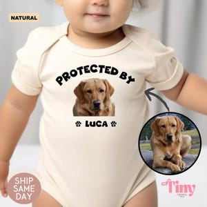 May include: Cream-colored baby bodysuit with the text "PROTECTED BY" above a photo of a golden Labrador Retriever. The name "LUCA" is below the photo, with paw prints on either side. A second photo of the dog is on the right.